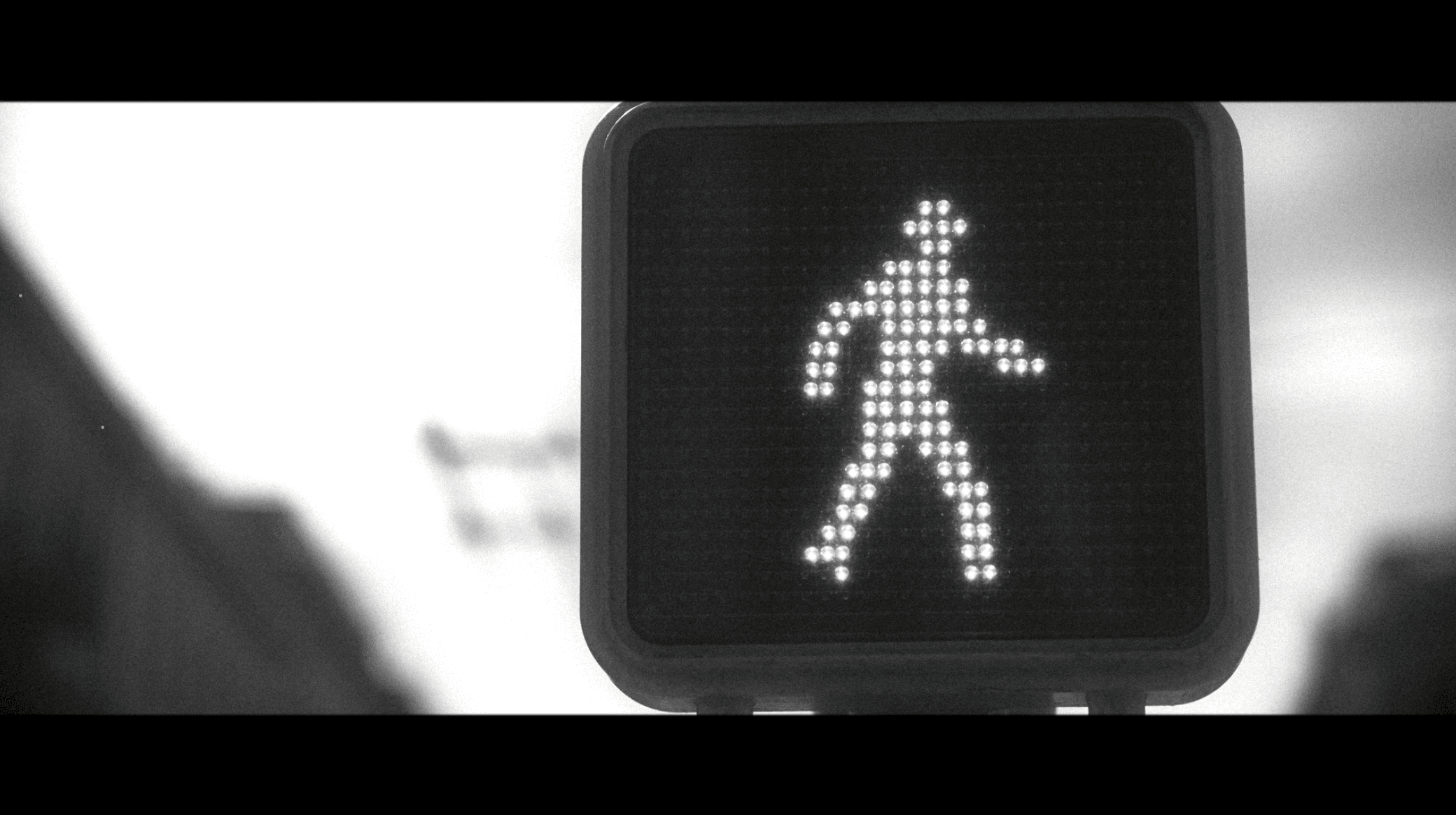 Graded still: a walk signal lit against a blown-out empty street